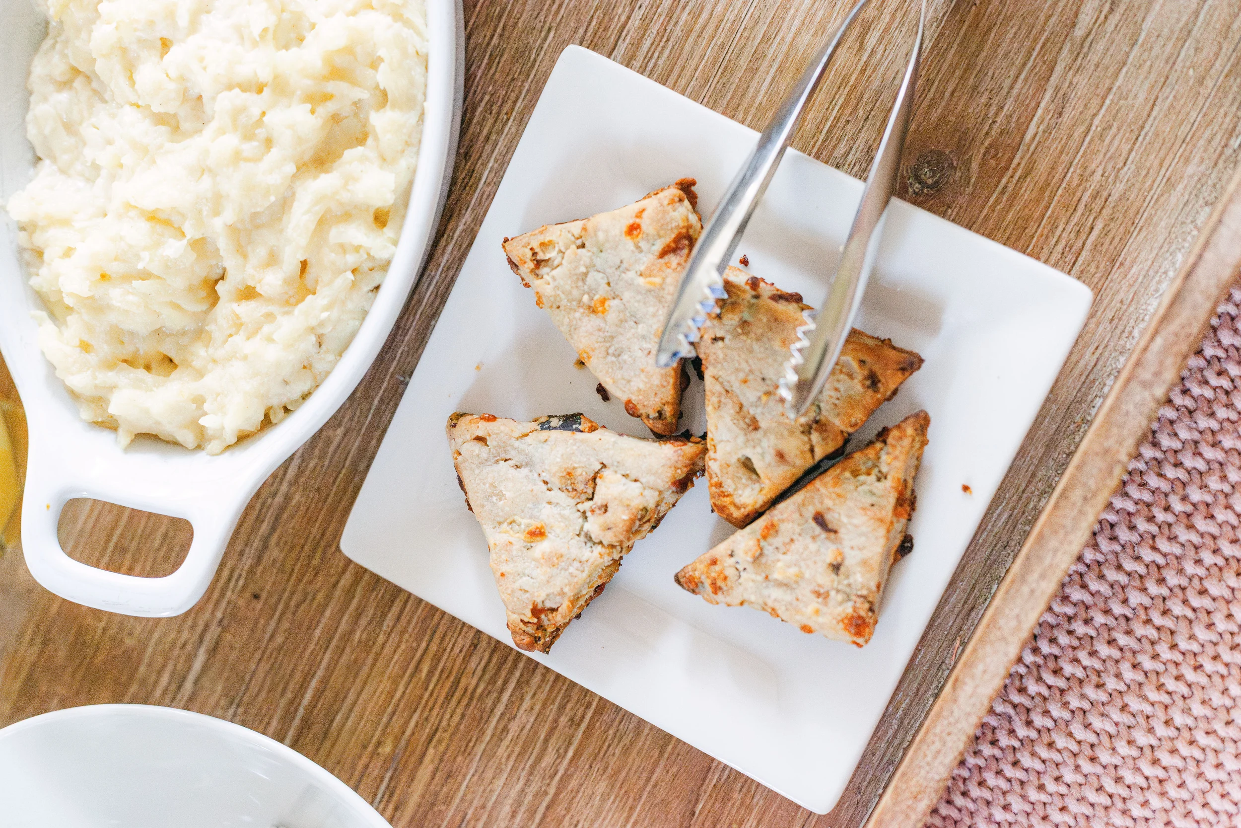 Pictured are four scones on a white plate with tongs to serve. There is also a white bowl with handles that contains grits. 